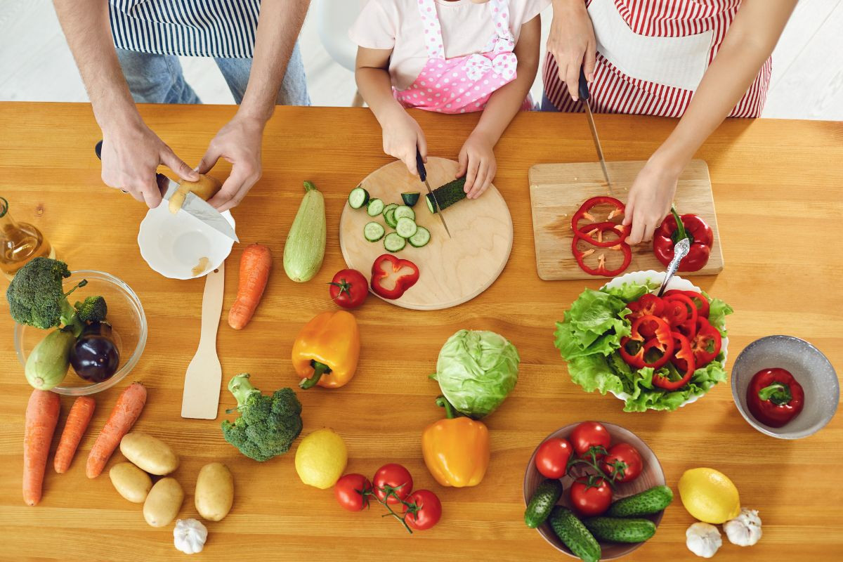 A family slicing various fresh vegetables together at a wooden table.